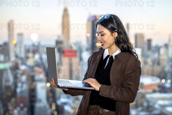 Young asian businesswoman using a laptop while standing against a blurred manhattan cityscape, representing urban professionalism, global business, and modern technology