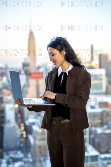Young businesswoman standing by a window, focused on her laptop while working in an urban setting with a new york city skyscraper backdrop, representing business ambition and success