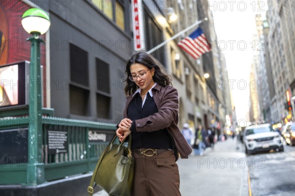 Young professional woman in a brown jacket checks her watch by a manhattan subway entrance, poised and punctual before starting her commute through the financial district