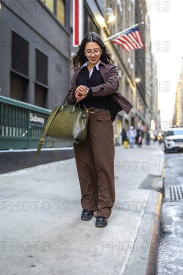 Young businesswoman walking urban street checking wrist watch while holding a handbag, representing punctuality, busy executive lifestyle, and urban commuting