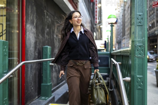 Young businesswoman with glasses and a professional bag emerging from a subway station entrance, confidently walking on a city street in new york, embodying urban success
