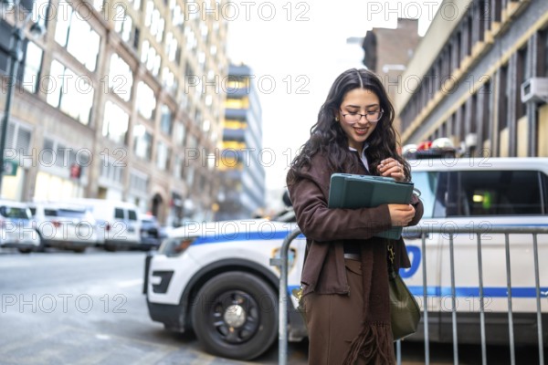Young executive woman is holding a modern tablet and smiling, standing outdoors in a bustling city street with buildings and a police vehicle in the background