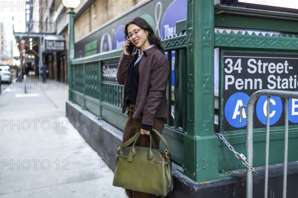 Young businesswoman in a brown jacket and glasses stands on a manhattan sidewalk near penn station, smiling while making a call on her mobile, commuting confidently through the city