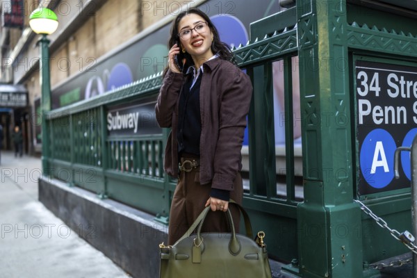 Young businesswoman smiling, talking on a mobile phone, and carrying a handbag while standing near a subway entrance in manhattan, new york, representing urban life and commuting
