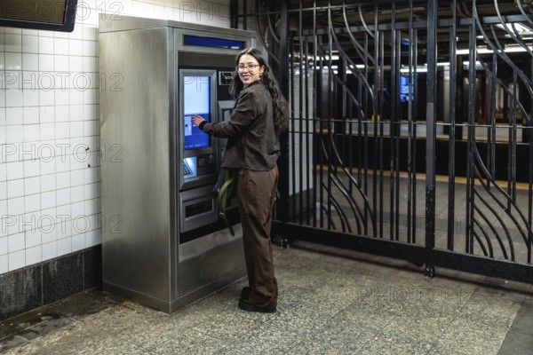 Young businesswoman with glasses and dark wavy hair smiling while interacting with a ticket machine at a subway station, preparing for her daily commute in manhattan