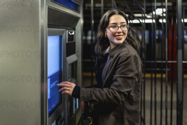 Young businesswoman with glasses and dark coat smiling, looking over her shoulder while interacting with a subway ticket machine in a busy urban station