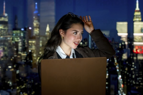 Young businesswoman working on laptop, thinking and looking away from a high rise office window at the illuminated manhattan skyline and city lights at night