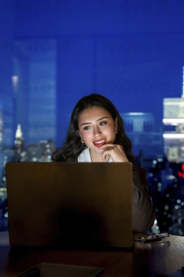 Young businesswoman working late on a laptop inside a modern manhattan office, illuminated by the screen light with the urban skyline of new york city visible through the window at night
