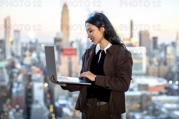 Young businesswoman working on a laptop, standing with a new york city skyline as a blurred background, representing modern business, global communication, and urban professional life