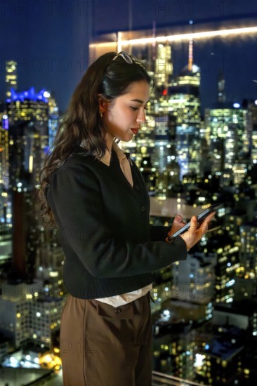 Young businesswoman standing by a window, engrossed in her smartphone, with the illuminated manhattan skyline serving as a dynamic backdrop, symbolizing connectivity and urban professional life