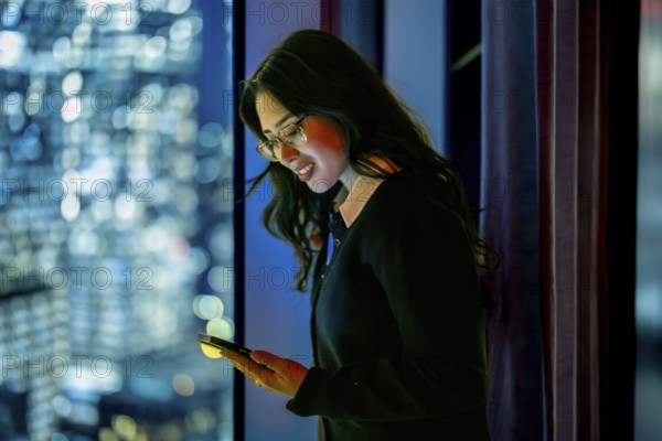 Young businesswoman standing by a skyscraper window at night, focusing on her mobile phone screen bathed in its glow, representing global communication and urban success