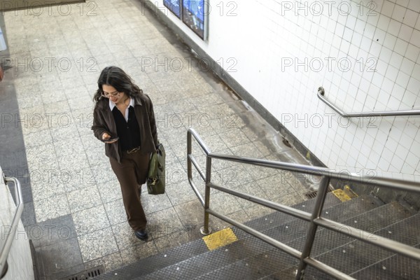 Young businesswoman walking down subway station stairs, checking her smartphone while holding a briefcase, representing urban professional life and daily commute