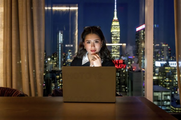 Young businesswoman with glasses on her head, focused on her laptop in a high rise at night, overlooking the illuminated new york city skyline and empire state building