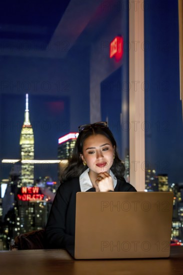 Young asian businesswoman focusing on her laptop screen, working late from an office with the illuminated manhattan skyline, including the iconic empire state building, visible outside the window