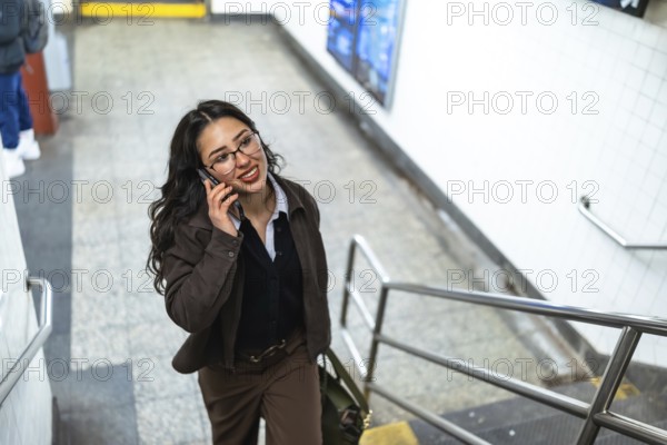 Young businesswoman with glasses and a professional outfit talking on a mobile phone while confidently walking up stairs in a bustling manhattan subway station
