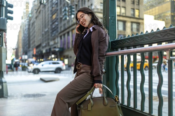 Young businesswoman smiling on a phone call, leaning against a green subway railing on a busy manhattan street, conveying modern urban commuter lifestyle and confidence