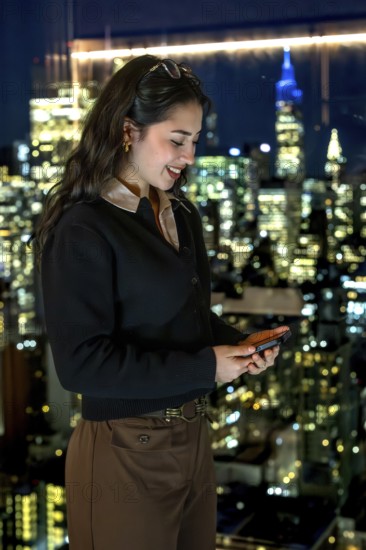 Young businesswoman smiling, looking at her smartphone while standing by a window with a stunning cityscape view of manhattan at night, reflecting success and modern communication