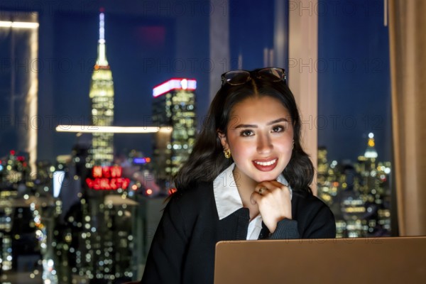 Young businesswoman smiling at camera, sitting in front of a window with a blurred night view of the illuminated new york city skyline and the empire state building