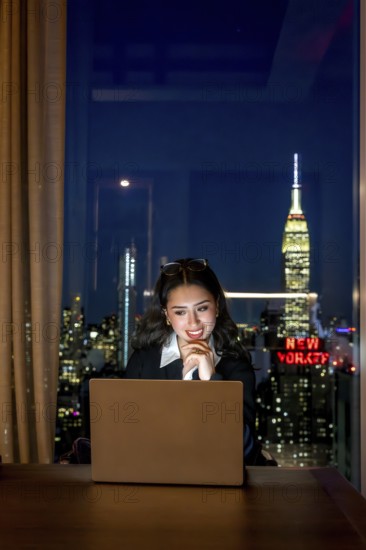 Young businesswoman observing laptop screen with a thoughtful smile, working late in a high rise office overlooking the illuminated manhattan cityscape and iconic buildings