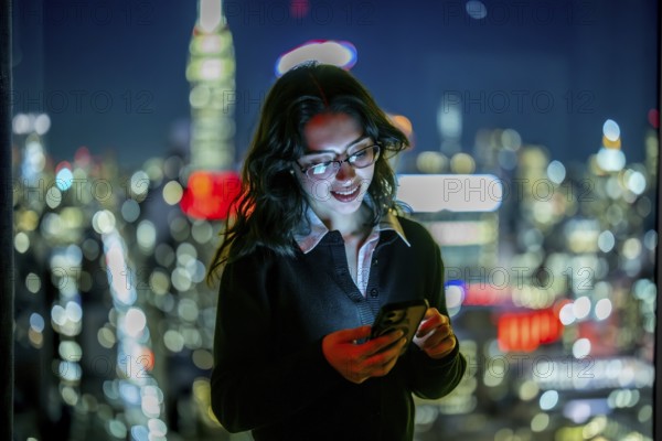 Young businesswoman smiling, reading a message on her glowing smartphone, standing by a window with a blurred manhattan city skyline at night, representing modern communication and urban life
