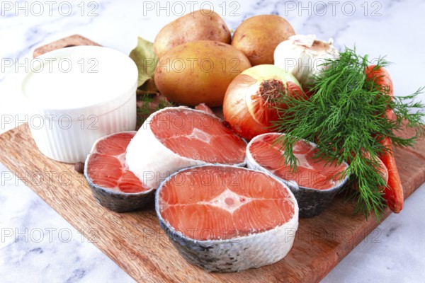 Potatoes, salmon, garlic, onion, and herbs are placed on a wooden cutting board. These ingredients are ready for meal preparation in a kitchen