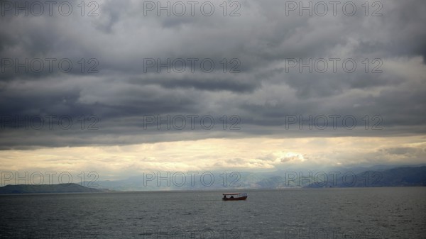Small boat on calm sea under cloudy sky surrounded by mountains in the background, Lake Ohrid, North Macedonia