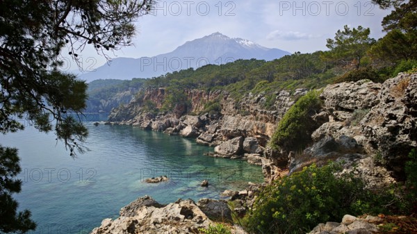 A rocky coast with clear blue water and green vegetation, with a snow-capped mountain rising in the background, view of Tahtali, Lycia, Antalya, Turkey