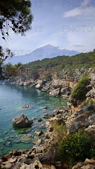 Rocky coast with clear blue water and view of snow-capped mountains in the background, view of Tahtali on the Lycian Way, Antalya, Lycia, Turkey