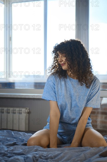 A young woman is sitting on a bed in a simple room. Bright daylight comes through the window, highlighting her curly hair and relaxed pose. The bedding looks soft, and the walls have a warm tone