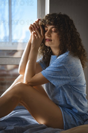 Dreamy woman with curly hair sits on a bed, resting hands together in front of their face. The soft morning light comes through the window, creating a warm atmosphere in the room