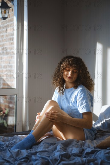 Dreamy woman is sitting on a bed near a window. She is looking down while touching her leg softly. Sunlight is shining through the window, creating a warm atmosphere in the room