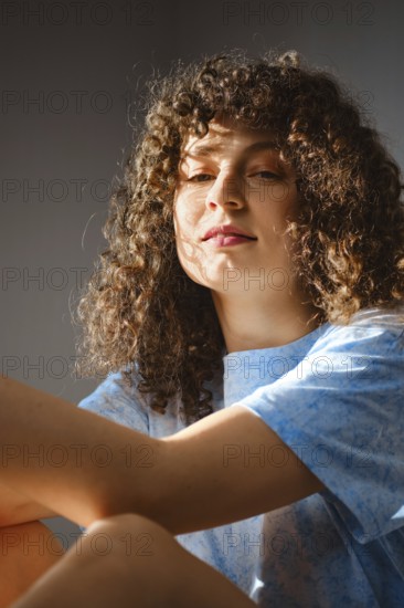 A young woman sits in a room, allowing sunlight to shine on her. She has curly hair and wears a blue shirt. The warm light creates a soft atmosphere as she looks contemplatively towards the light
