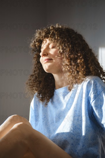 A woman sits relaxed by a window with sunlight on her face. She has curly hair and wears a blue shirt. The light creates soft shadows around her as she enjoys a quiet moment indoors in the afternoon