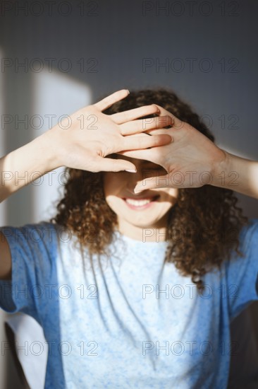 A woman stands in a well-lit room, smiling and covering her face with both hands to block sunlight. She has curly hair and wears a blue shirt. The light creates shadows around her