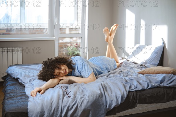 A young woman lies on her stomach on a bed, wearing blue pajamas. Sunlight comes through the window and shine her feet. Plants are visible outside. The room feels bright and airy