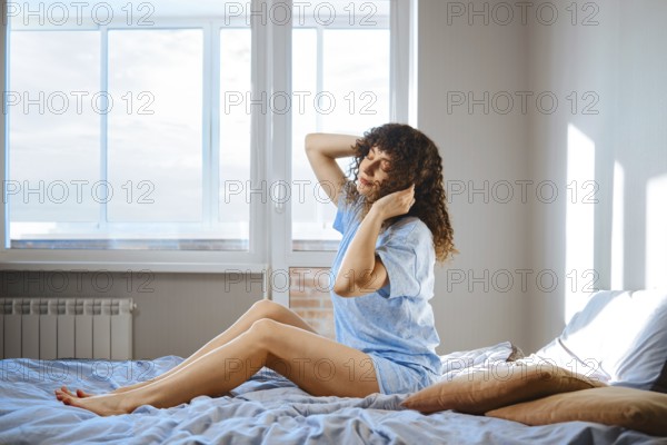 A woman sits on her bed in a bright room. She stretches her arms and turns her head as sunlight comes through the window. The room has a cozy feel with soft bedding and pillows