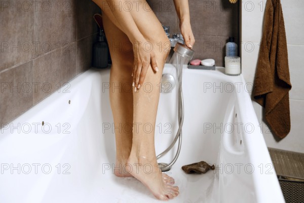 A woman stands in a bathtub washing her legs with a shower head. The bathroom has tiled walls, toiletries on display, and a towel hanging nearby. Water runs down her legs