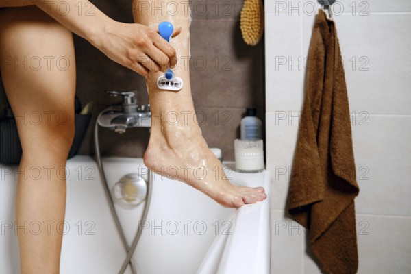 Unrecognizable woman is standing in a bathtub with one leg raised while using a razor to shave. Typical morning skincare routine in a home bathroom