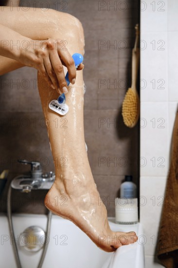Unrecognizable woman put her leg on the edge of a bathtub and using a razor to shave her leg. Shaving cream covers the skin. The bathroom has a tidy setup with bath products