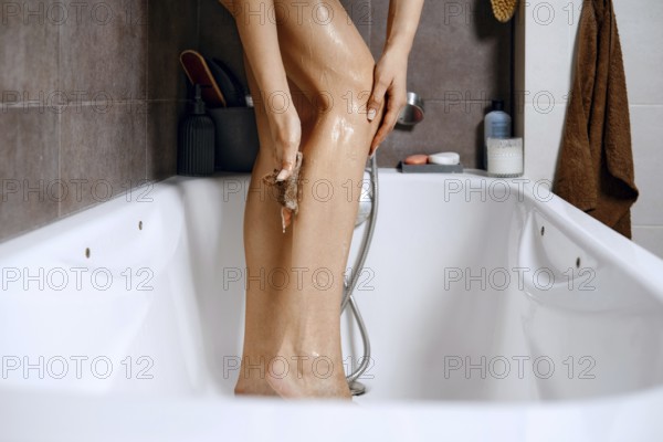 A woman stands in a bathtub. She uses a scrubbing tool to wash her leg. The bathroom has tiles, a shower head, and bath products nearby. Water and soap are visible as she cleans