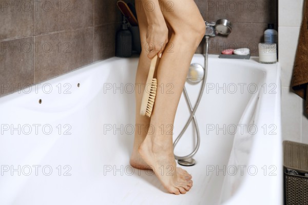 Woman stands in a bathtub during a personal care session. She hold a wooden massage brush in one hand and use it on her leg. Items are placed nearby for convenience