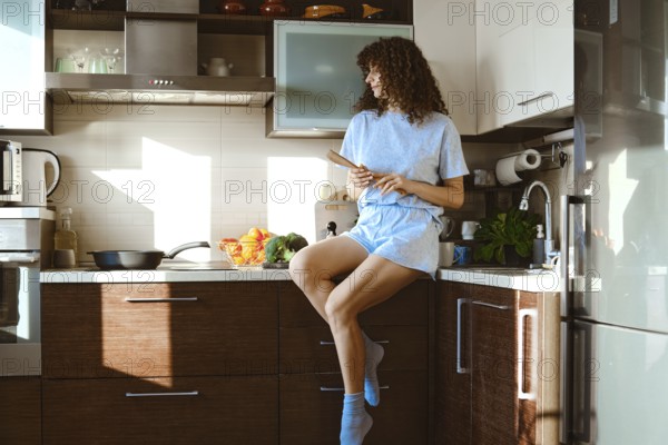A woman sits on a kitchen counter in a modern home. She holds a spatula and looks at frying pan. The sunlight comes in through the window. The scene shows a busy kitchen with various items