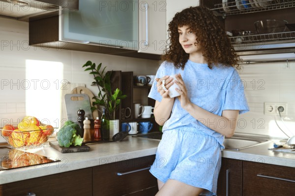 Smiling woman stands in a kitchen, sipping coffee from a mug. Fresh fruits and vegetables are on the counter. Bright light comes through the window, creating a warm atmosphere in the room