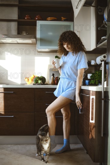 A woman is in the kitchen wearing a light blue outfit. She holds a drink in one hand while her cat is by her feet. Sunlight fills the space as she begins her morning routine