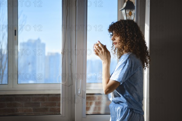 A woman stands by an opened for ventilation balcony door in the morning, looking outside while holding a cup. The sunlight shines in, and a city skyline is visible. She is wearing casual clothing and seems lost in thought
