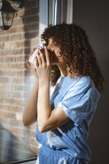 A woman stands by a window and holds a warm drink close to her face. Sunlight shines through the window and illuminates her curly hair. She seems to enjoy the moment in her home during morning time