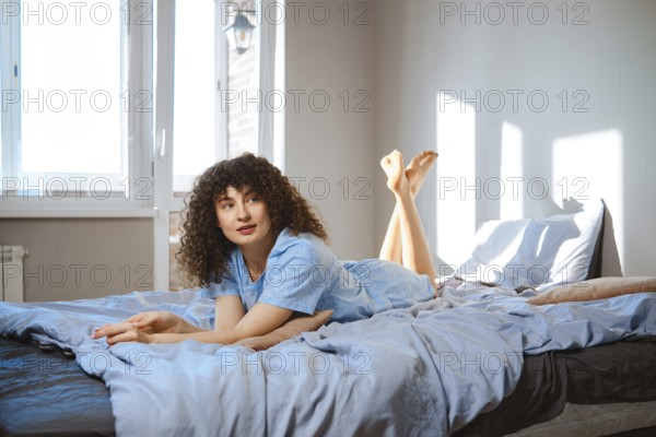 A woman with curly hair lays on a bed with light blue sheets. She looks to the ceiling while sunlight enters the room