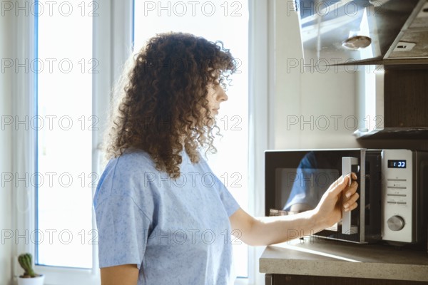 A woman stands by a microwave in a kitchen. She is preparing to heat food. Sunlight enters through the window. The kitchen is tidy with simple decor. There is a plant in the background