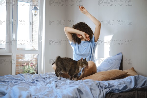 Young woman is sitting on a bed and stretches her arms in a room with light coming through a window. The cat is walking around the bed. This scene shows a morning routine in a cozy space