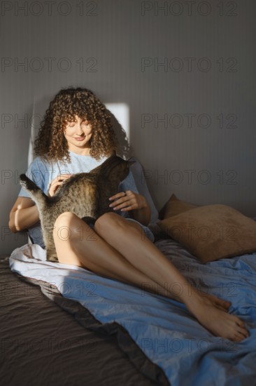A woman sits on a bed in a cozy room during morning light. She gently pets a cat sitting on her lap. The setting shows a relaxed atmosphere with soft bedding and warm colors